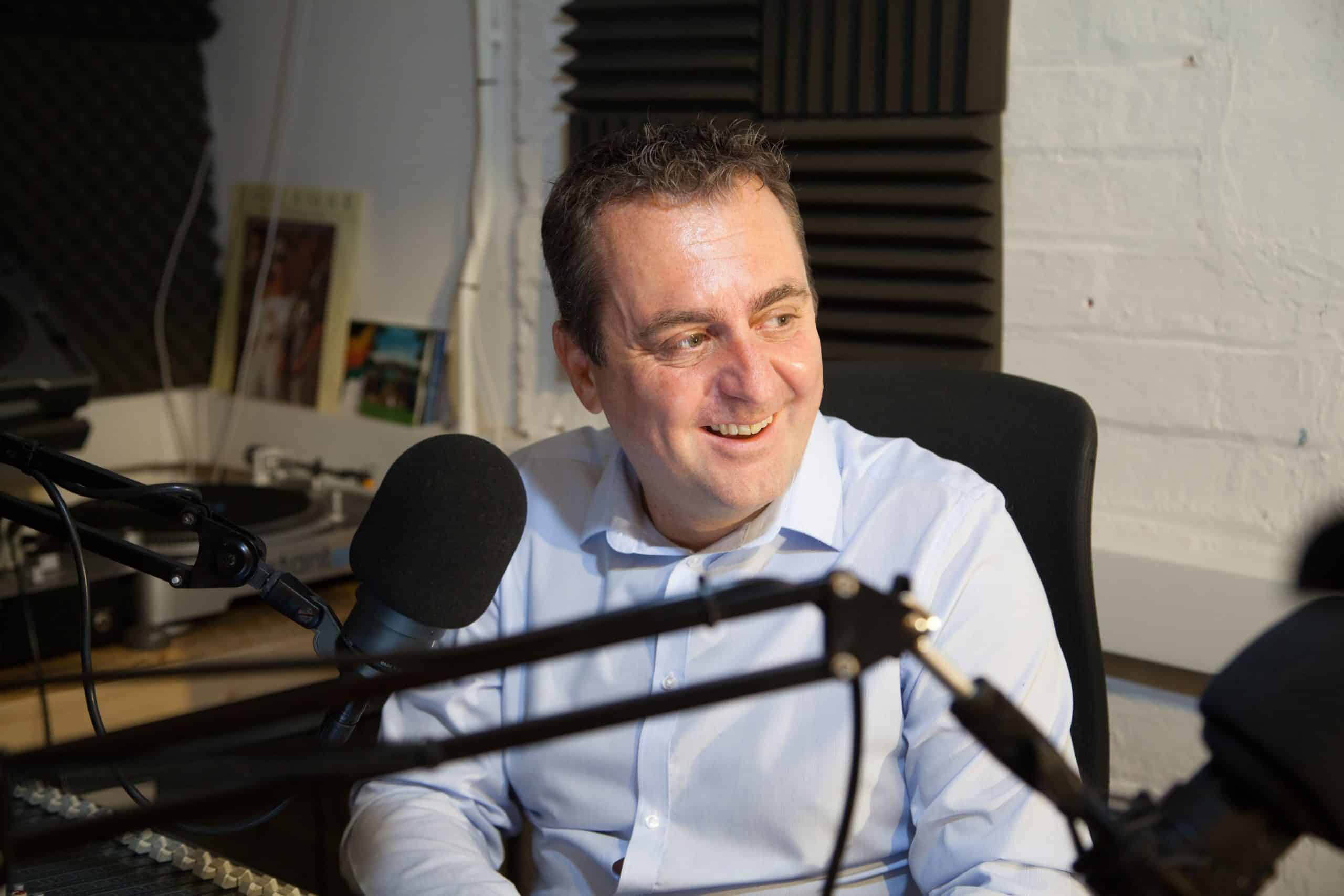 Neil C. Hughes i ljusblå skjorta i ljudstudio med mikrofon, glad och fokuserad på samtal i sin podcast.