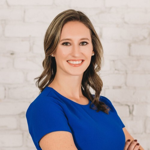 Ashley Rose stands with her arms crossed and her shoulders angled at three-quarters from the camera. Her face is turned to face the camera head-on. Rose is smiling and wearing a royal blue boatneck blouse.