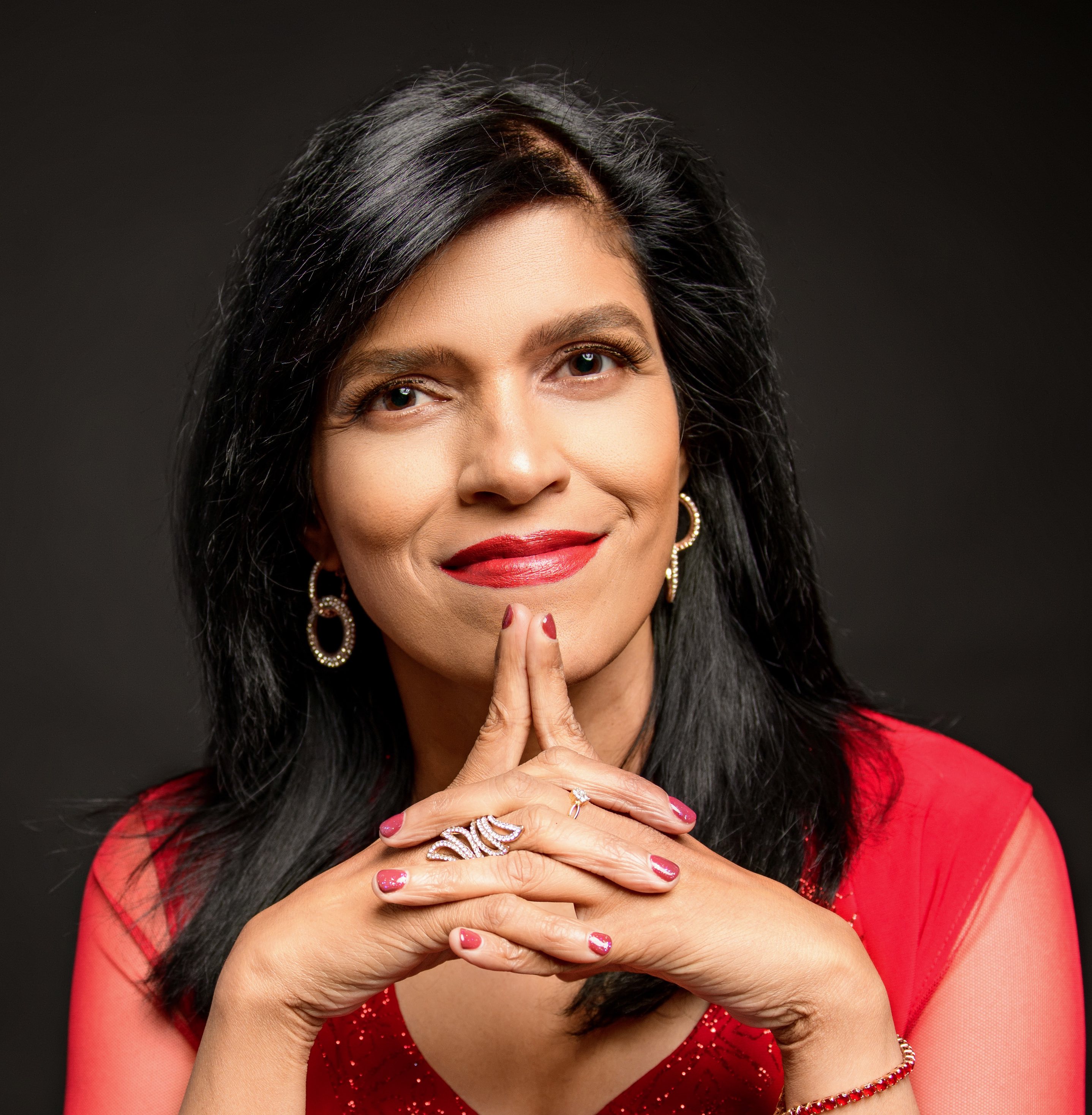 Beena Ammanath faces directly toward the camera wearing a red blouse and gold hoop earrings. She clasps her hands just beneath her chin.