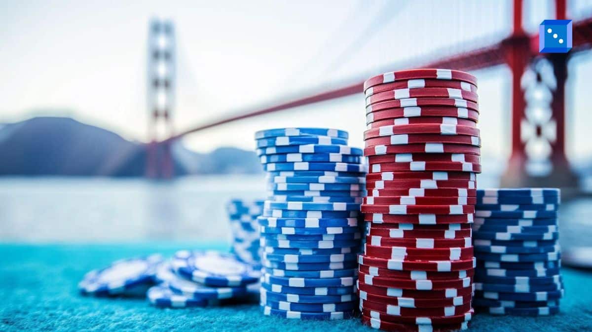 Stacks of red and blue poker chips sit on a vibrant blue felt table with the Golden Gate Bridge in the background. The iconic bridge is slightly blurred, creating a dynamic contrast between the sharp focus on the gambling chips and the famous San Francisco landmark. The scene evokes themes of risk, fortune, and high-stakes betting.