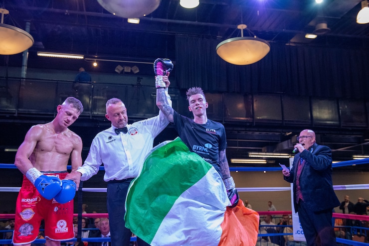 The referee is holding the wrestlers by their hands, raising Lee Gormley's hand up in the ring