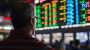 A man in a red-striped shirt and glasses closely observes a large electronic betting board displaying colorful odds and statistics at a sportsbook.”