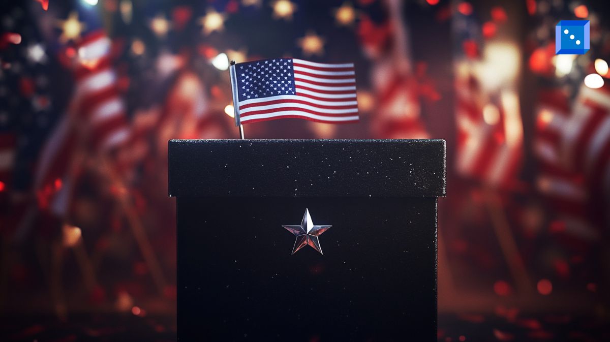 A black ballot box adorned with a silver star and a small American flag sits in the foreground, symbolizing democracy and elections. The background is filled with blurred American flags and festive red, white, and blue confetti, creating a patriotic atmosphere.