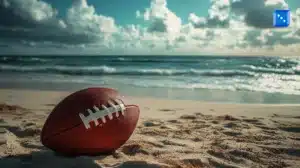 An American football rests on a sandy beach with ocean waves rolling in the background. The sky is partly cloudy, with sunlight reflecting off the water. A small blue icon with white dots is positioned in the upper right corner.