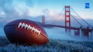A close-up of an American football resting on frost-covered grass with the Golden Gate Bridge in the background, partially covered in mist. The sky displays a blend of blue and pink hues, adding depth to the image. A small blue icon with white dots is located in the upper right corner.