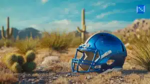 A shiny blue American football helmet rests on the desert ground among cacti and dry grass. The background features towering saguaro cacti, mountains, and a bright blue sky with scattered clouds. A small blue icon with white dots is positioned in the upper right corner.”