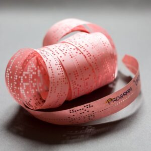 A roll of pink punch tape with perforated dots, featuring the word "PROGRAM" printed on the side, rests on a gray surface.