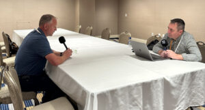 Two men sit at a long white table in a conference room, one holding a microphone, the other with a laptop open in front of him.