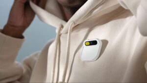 A person wearing a beige hoodie with a compact, white device featuring a camera and yellow button clipped to the chest area.