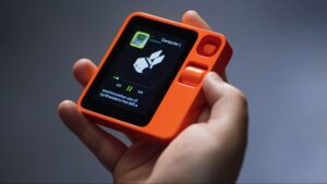 A person holding a vibrant orange Rabbit R1 featuring a screen displaying playback details of a song.