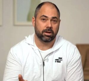 A person wearing a white zip-up hoodie sits against a neutral background, gesturing with hands as if engaged in conversation.