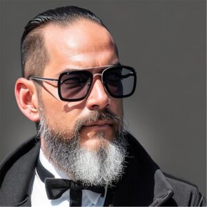 A well-dressed individual with a gray beard and stylish glasses, wearing a black tuxedo with a bow tie against a neutral background.