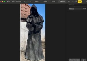 A tall, cloaked figure with a hood stands silently, hands clasped, against a backdrop of blue sky and rooftops.