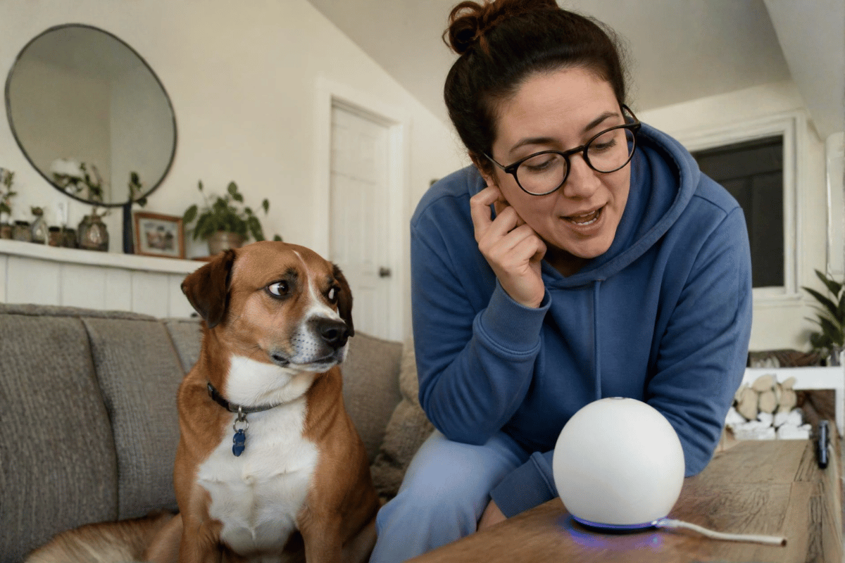 Woman speaking to a smart speaker while her dog gives it a suspicious side-eye, illustrating the rise of AI companion devices and voice assistants in the smart home. OpenAI’s Portable AI Speaker Could Change the Smart Home