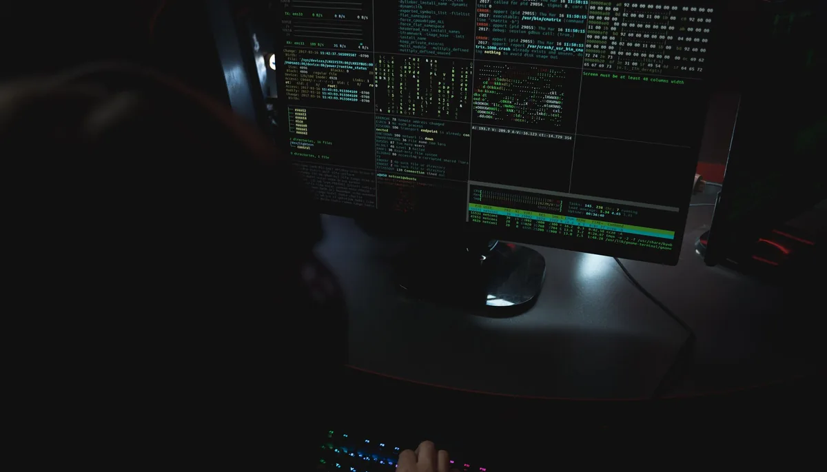 A person typing at a computer with monitors showing code and system data in a dark room.